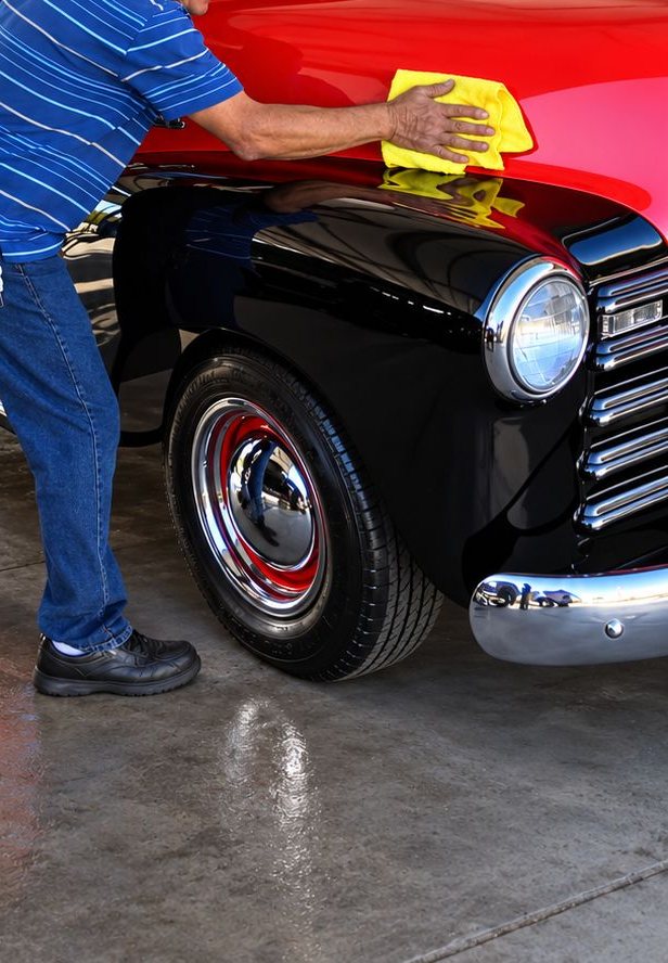 A hand polishing the red hood of a classic pickup with a yellow microfiber towel, above a glossy black fender, chrome headlight, grille and bumper, and a red wheel with a chrome hubcap