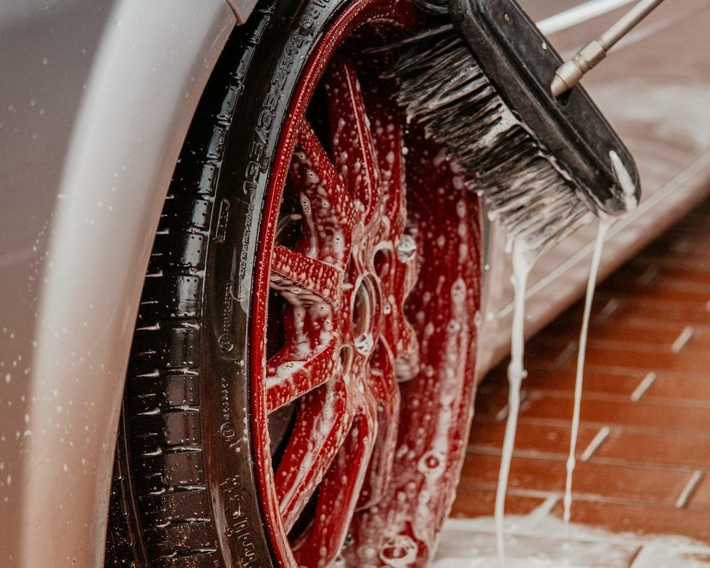 A long-handle brush scrubbing soapy foam over a red wheel and tire